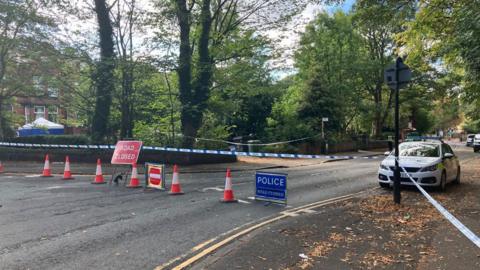 A street is taped off by police, with a blue forensic tent seen in the distance near a house. A police car is parked on the right. The street has trees on both sides, with some leaves on the ground. 