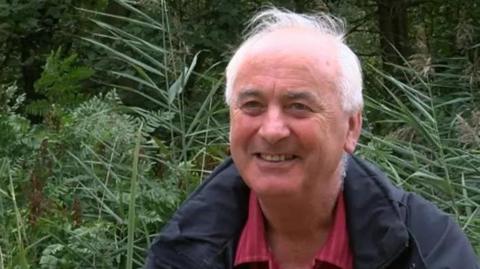 Prof Alastair Fitter wearing a red shirt with white stripes and a blue waterproof coat photographed in front of bracken and reeds at Askham Bog