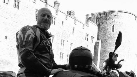 Pawel Dlugosz is sat on a motorcycle in front of a castle. The photo is in black and white. He is bald and has a beard, and is wearing a leather jacket and trousers. 