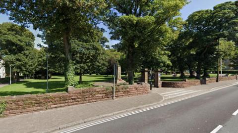 A stone wall lines a park with green fields and trees. A road runs alongside it, to the right. It is a sunny day.