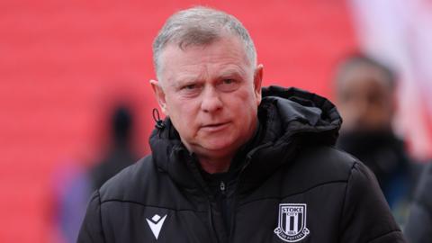 Stoke city manager Mark Robins looks firmly ahead with a focused expression on his face. he wears a black puffer coat with the stoke city badge on it.