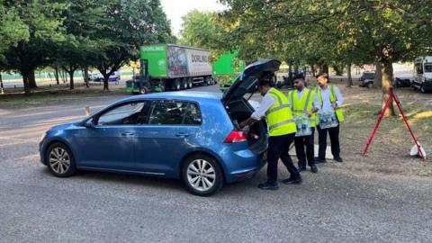 Three men load bottled water in the boot of a blue car. 
