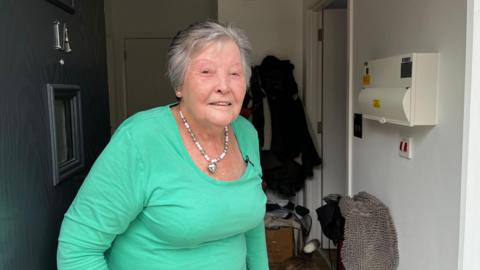 A grey-haired woman with green top stands at her front door