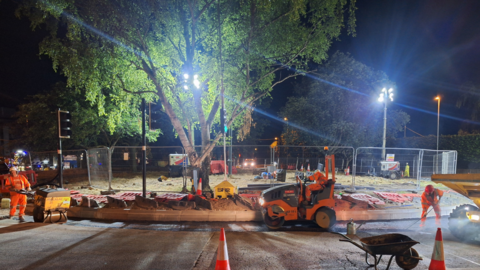 Construction work is taking place overnight. Bright flood lights have been put on the centre of a roundabout as workers in orange high vis outfits use machinery on the roads. There is a large tree in the centre of the roundabout. 
