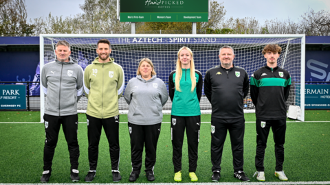 Representatives of Guernsey FC’s teams. Left to right: Tony Vance, Ross
Allen, Anna Gauvain, Heidi Dawber, Leon Meakin and Tyrese Kelly. They are wearing Guernsey FC football clothing and are all facing forward with their hands behind their back. The goal it behind them. The pitch is green. Behind the goal is a blue wall. Behind it are the tops of trees.
