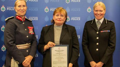 Three women in a row. The woman in the middle is holding an award in a frame and has short brown hair with blonde highlights. She is wearing a dark jacket. The woman on the left is wearing a dark uniform and has medals and has her blonde hair tied back. The third woman has her blonde hair tied back and is wearing a black police uniform and also has medals on her lapel. 