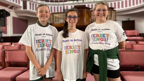 Three girls are smiling and wearing T-shirts saying Music in Action and Jersey Sings! They stand in front of chairs in St Helier Methodist Centre. 