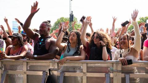 Crowd of festivalgoers standing behind a metal safety barrier at an outdoor music event, many with their hands raised in the air. A group at the front includes several young adults leaning on the barrier, some holding drinks and mobile phones, with trees and stage equipment visible in the background under bright daylight.