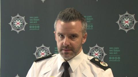 An officer is talking into mics. He's wearing a white shirt with black lapels.