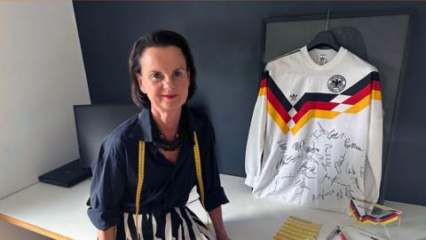 A woman sits next to a 1990 West Germany shirt hung up. She's wearing a dark blue shirt. 