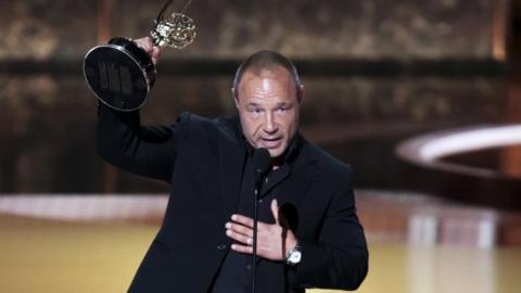 Stephen Graham has short hair and is wearing a black suit and black shirt. He is holding an Emmy award aloft with his right hand and had his left hand on his chest.