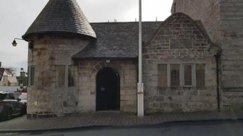The outside of the former police station, which is a small stone-built structure with a turret-shaped section on the left and a flagpole in front of it.