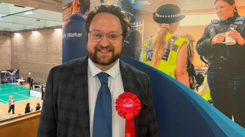 Matt Storey, who has swept back dark hair and a beard. He's wearing glasses with clear frames, a grey suit and a red Labour rosette.