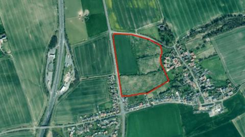 An aerial map of the proposed site highlighted by a red boundary. The boundary is surrounded by green fields with a small settlement to the right. To the left there is a major road.