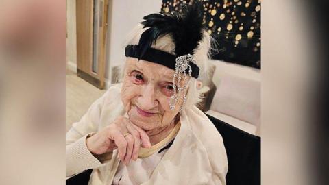 Head and shoulder shot of an 99-year-old woman, Maisie Dalziel. She is smiling, holding her right hand up to her chin, and wearing a cream coloured cardigan over a light coloured top. Her hair is white and on her head is a black headband, with a feather and a diamante attachment.