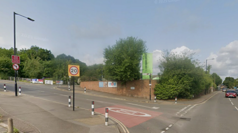 Street view of a junction in a suburban area. The road splits into two directions: one going uphill to the left with a 20 mph speed limit sign and bollards, and another continuing straight ahead marked as B6088. A brick wall with posters and a green sign is visible at the corner, surrounded by trees and greenery.