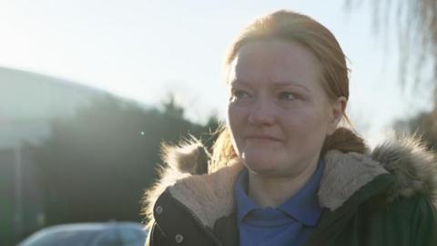 A tearful woman in a blue jumper, blue polo shirt and olive coat looks off to the right.