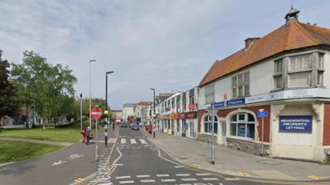 A Google Street View image of Alexandra Parade. To the left is a long green space with trees, running down the middle of the road, while a parade of shops and businesses is on the right. The road ahead is one-way, with a pedestrian crossing directly in front. 