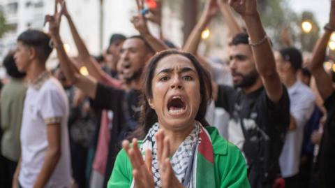 A protester looks at the camera as she chants a slogan. She is wearing a green top and is clapping her hands