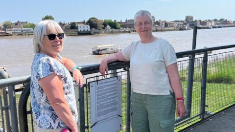 Alison Fletcher and Karen Champion stand in front of the Ferry which is moored in The Great Ouse river. 
Alison is wearing a white and blue top and dark sunglasses and Karen is wearing a white top and green shorts. 