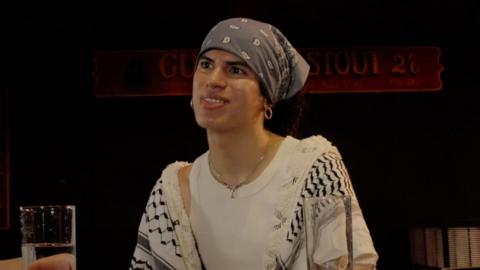 Jacob Alon is wearing a white vest top and black and white scarf over their shoulders. They have a light blue bandana tied around their head. They are looking off to the left of the image.