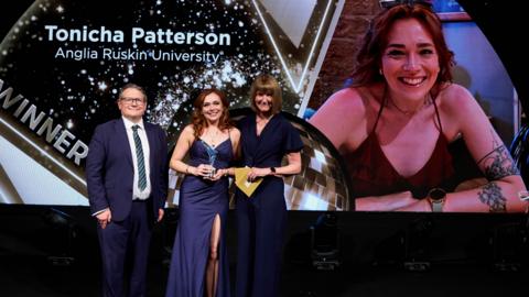 Tonicha, a woman, wearing a blue long dress standing on a stage being presented with an award - a second woman in a dark dress and a man in a blue suit are standing on either side of her.