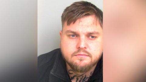 A mugshot of man with short light brown hair and a short beard. He has a tattoo on his neck.