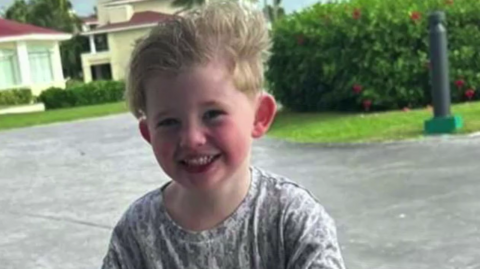 Peter Vaughan is outside in front of houses and lawns. He has blond hair and is wearing a grey T-shirt and is smiling at the camera, looking happy. His hair is standing up and his face is flushed.