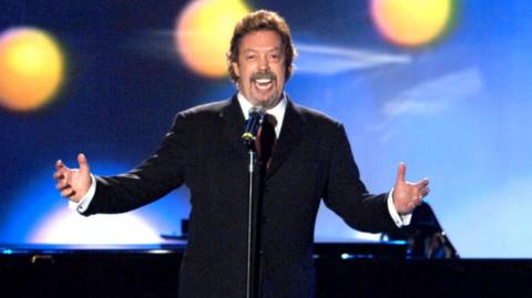 Tim Curry in a black suit with arms outstretched sings into a microphone