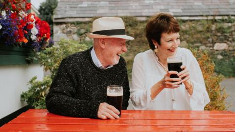 A man and a woman sit at an orange garden bench outside a pub. They are laughing and both holding pints.