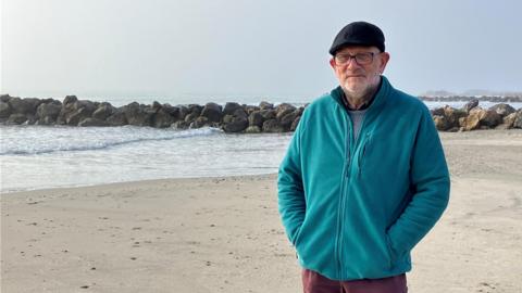 Mick is standing on a beach facing the camera. He is wearing glasses and a flat cap. He is standing with his hands in his pocket and is wearing a teal fleece and burgundy trousers.  