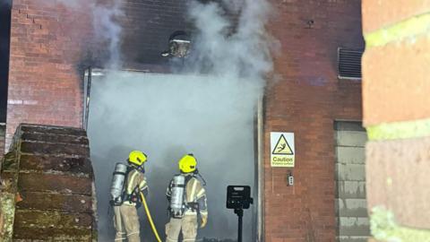 Two firefighters, standing outside a building, in full uniform, wearing helmets and breathing apparatus. There is smoke filling the doorway. There is a large red brick building with a sign to the right.