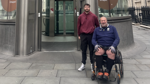 Chris during the London leg of the challenge. He is with his son and they are smiling at the camera. They are on a pavement with an entrance to a station behind them. 