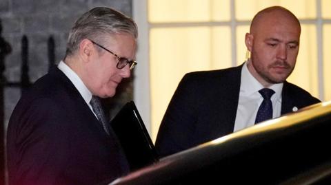 Keir Starmer ducks as he is about to get into the back seat of a black car while holding a folder, next to another man in a suit who holds the door open for him, taken outside Downing Street on 9 February.