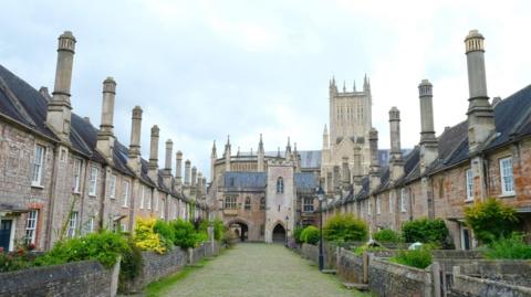 Vicars' Close with Wells Cathedral in the background, the close is paved with grey cobbles with grass growing through them. On either side are rows of terraced houses with large chimneys extending from the roofs. 