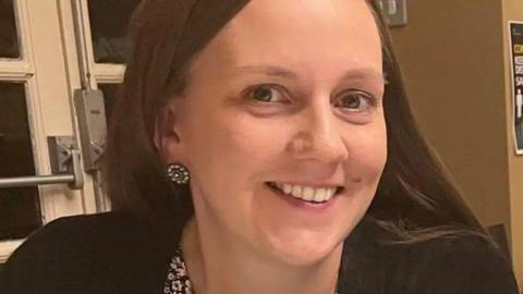 Jessica Dominy, pictured close-up. She is smiling at the camera, and is wearing large stud earrings, a black cardigan and a flower top. She has long dark hair. 