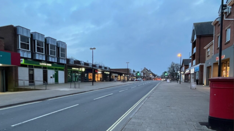 A road with wide pavements on either side, lined with shops.