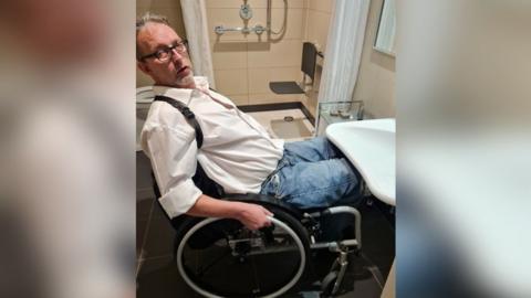 Rohan Tarry is in his wheelchair trying to access a sink but he cannot get close enough to it to reach it. He is looking at the camera.  Behind him is a bath with a disabled seating platform over it
