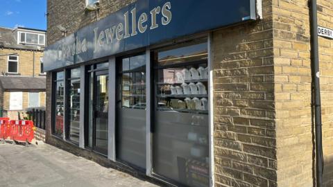 The outside of a jewellers, with the business advertising the name of the business above the door. The pavement in front has some red hoardings indicating roadworks. 