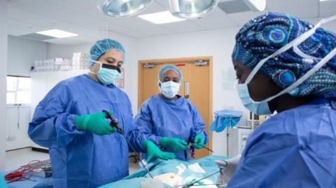 Students in blue gowns, wearing surgical gloves, masks and scrub caps practicing surgery.