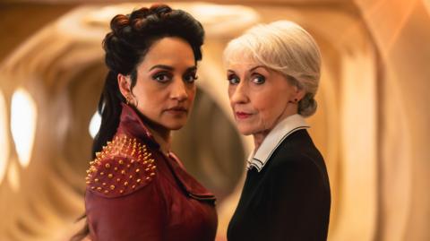 Archie Panjabi and Anita Dobson stand facing each other looking at the camera in a white room with rounded walls. Archie is wearing a red leather jacket with gold studded shoulders, and Anita is wearing a black sleeved dress with a white collar
