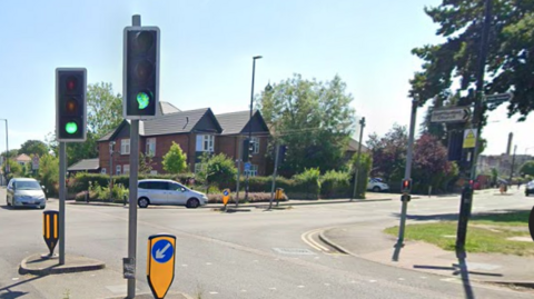 A wide road junction with traffic lights and a pedestrian crossing across Oatlands Drive. Stoke Poges Lane is to the right in the background.