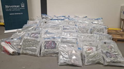 A photo of a large pile of clear plastic bags containing suspected herbal cannabis and cannabis resin. The bags have been placed on a floor next to a wooden pallet and a navy blue/dark green branded Irish Revenue sign which contains the organisation's name in Irish and its website address.