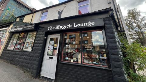 A street-level view of a small shop called The Magick Lounge. The building has a black-painted lower facade with a white door and large front window displaying various items. Above the shop sign, the upper part of the building is light-coloured with two windows and a mural of a silhouetted magician holding a wand.