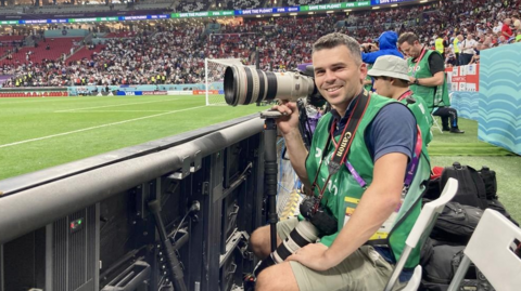 A man in a green bib over a blue polo shirt is sat at the side of a football pitch in a stadium. He is holding a large camera in one hand