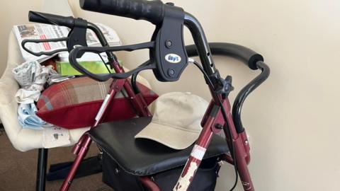 A zimmerframe is placed in the corner of a care home. The zimmerframe has a maroon colour and has a beige cap on the seat. There is a seat next to it with a newspaper.