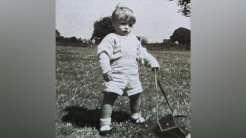 A black and white image of a toddler. The boy is playing outside. He is wearing dungarees with a long-sleeved top underneath, and socks and dark shoes.