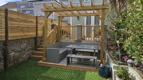 One of the restored gardens to the rear of a house. A wooden pergola sits on grass and holds a wicker-style outdoor dining set comprising an L-shaped sofa, a bench and a glass table. There is a new section of wooden fencing on one side and flowering shrubs in raised beds on the other. 