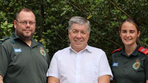 Gary Pendleton is wearing a short sleeved white shirt and is standing between the two ambulance crew members who saved him after a heart attack. He is smiling and has greying hair. Paul Arkle and Annie O'Sullivan are stood to either side of him in their dark green uniforms.
