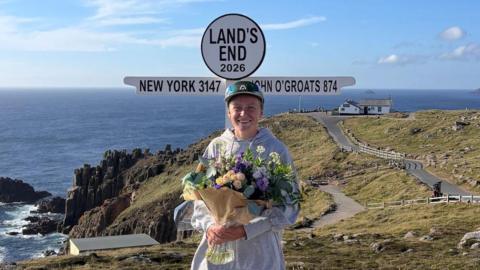 A picture of Ella Fryer holding a bouquet of flowers in front of a Land's End sign